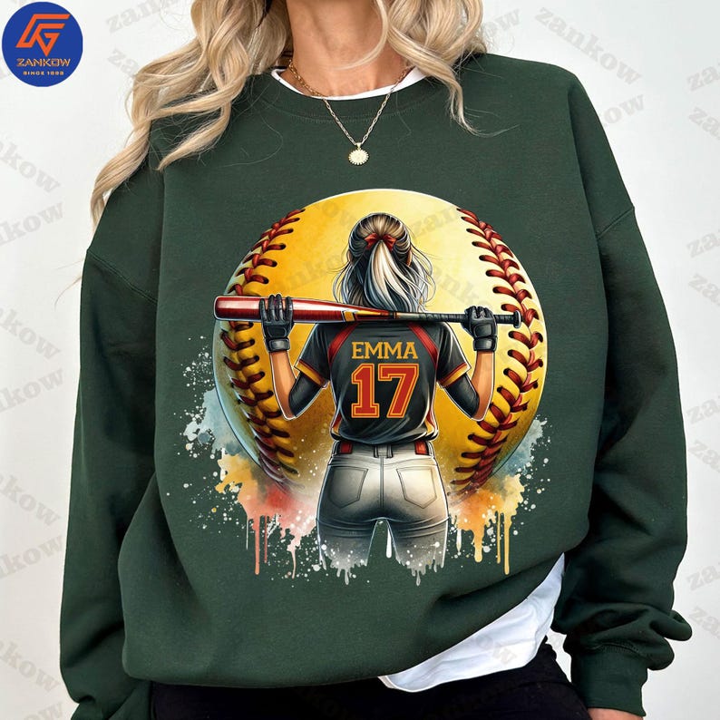 K&ouml;nnte beinhalten: Gr&uuml;nes Sweatshirt mit einem Aquarell-Design einer Softballspielerin, die einen Schl&auml;ger h&auml;lt. Die Spielerin tr&auml;gt ein schwarz-rotes Trikot mit dem Namen "EMMA" und der Nummer "17" auf dem R&uuml;cken.