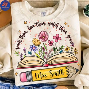 May include: A beige sweatshirt with a design of an open book with flowers growing out of it. The book has a yellow pencil with the text "Mrs. Smith" on it. The text around the book reads "teach them love them watch them grow".