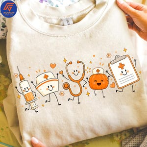 May include: A beige crewneck sweatshirt featuring a graphic print of five anthropomorphic medical items: a syringe, a nurse cap, a stethoscope, a pumpkin wearing a nurse cap, and a clipboard, all with smiling faces and orange accents.