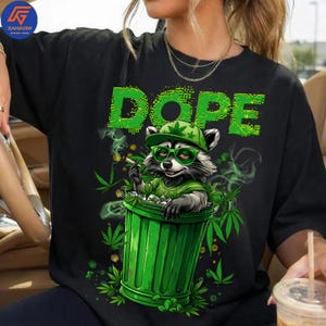 Puede incluir: Camiseta negra con un gráfico de un mapache en un cubo de basura verde, con gorro y gafas, fumando un cigarrillo. La palabra "DOPE" está en verde encima del mapache. Hojas de marihuana rodean al mapache.