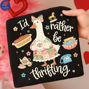 Puede incluir: Cojín cuadrado negro con un dibujo de un ganso de dibujos animados con un vestido de retazos y sosteniendo un plato. El texto "I'd rather be thrifting" está escrito en blanco. Elementos decorativos incluyen una tetera, cuencos y un teléfono.