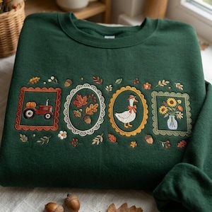 May include: A dark green crewneck sweatshirt featuring four embroidered rectangular and oval frames. The designs include a red tractor with a pumpkin, autumn leaves with an acorn, a white goose wearing a hat and bow, and a vase of sunflowers.