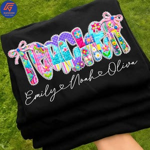 Pode incluir: Camisola preta com a palavra "TEACHER" em letras coloridas, estilo patchwork, decoradas com desenhos florais e estampados. Abaixo, o nome "Emily & Noah Oliva" está escrito em itálico. A camisola está dobrada.