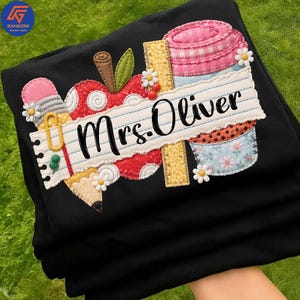 Op de afbeelding: Zwart T-shirt met een ontwerp met een lerarenthema. Het ontwerp bevat een appel, potlood, liniaal en een koffiekopje, met de naam "Mrs. Oliver" op een banner van gelijnd papier. Het ontwerp is versierd met kleine witte bloemen.