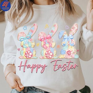 May include: White sweatshirt featuring three Easter bunny designs with floral patterns, bows, and patchwork details. The words "Happy Easter" are written in pink script below the bunnies. The design includes hearts and floral accents.