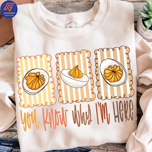Op de afbeelding: Crèmekleurige sweatshirt met een afbeelding van drie gevulde eieren in decoratieve kaders. De tekst "YOU KNOW WHY I'M HERE" staat eronder in oranje en bruine letters. Een casual kledingstuk met een voedselthema.