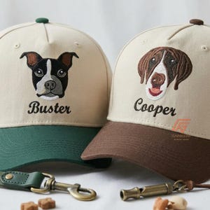 Puede incluir: Dos gorras de béisbol beige con retratos de perros bordados y nombres. La gorra izquierda tiene un perro blanco y negro llamado Buster con visera verde. La derecha tiene un perro marrón y blanco llamado Cooper con visera marrón y un logo naranja.