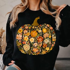 May include: Black sweatshirt featuring a floral pumpkin design. The pumpkin is composed of various flowers in shades of orange, yellow, and peach, with a green stem. The design is centered on the front of the sweatshirt.