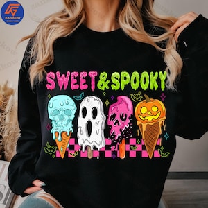 Op de afbeelding: Zwarte sweatshirt met de tekst "SWEET & SPOOKY" in roze en groen. De afbeelding toont vier ijsjes met spookachtige ontwerpen: een schedel, een spook, een roze schedel en een pompoen. De ijsjes staan op een roze en zwart geblokt patroon.