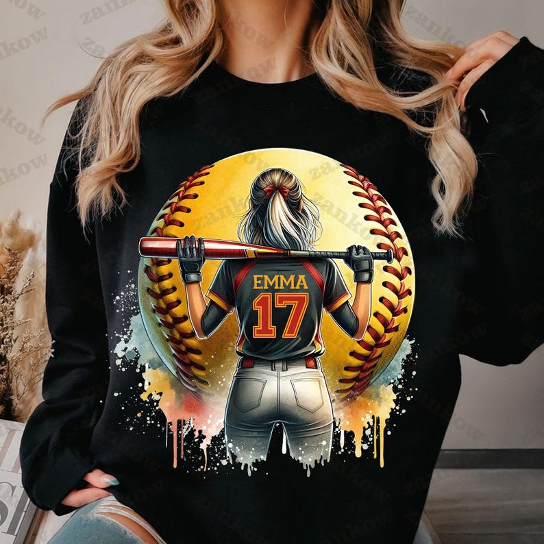 K&ouml;nnte beinhalten: Schwarzes Sweatshirt mit einem Grafikdruck einer Softballspielerin, die ein Trikot mit dem Namen "Emma" und der Nummer "17" tr&auml;gt. Die Spielerin h&auml;lt einen Schl&auml;ger und steht vor einem gelben Softball.