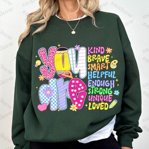 May include: Dark green sweatshirt with the words "You Are" in colorful, patterned letters. The words "Kind, Brave, Smart, Helpful, Enough, Strong, Unique, Loved" are also displayed in a playful font. A pencil and flower graphics are included.