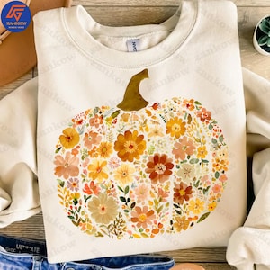 May include: Cream-colored sweatshirt featuring a floral pumpkin design. The pumpkin is composed of various flowers in shades of orange, yellow, and peach, with green leaves. The stem is brown. A cozy fall-themed garment.