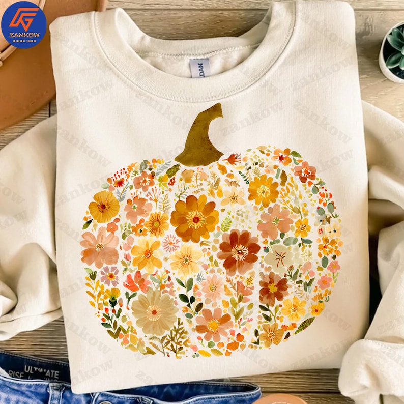 May include: Cream-colored sweatshirt featuring a floral pumpkin design. The pumpkin is composed of various flowers in shades of orange, yellow, and peach, with green leaves. The stem is brown. A cozy fall-themed garment.