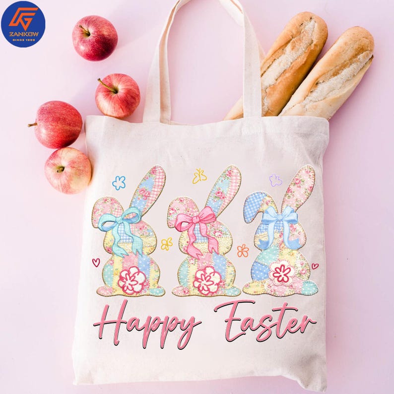 May include: A cream-colored tote bag featuring three patchwork bunny rabbits with floral and gingham patterns, each with a bow. The words "Happy Easter" are printed in pink. Apples and baguettes are also visible.