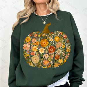 May include: Dark green sweatshirt featuring a floral pumpkin design. The pumpkin is composed of various flowers in shades of orange, yellow, and peach, with a brown stem. The sweatshirt has a white crew neck.