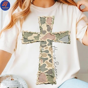 May include: A cream-coloured t-shirt with a large, textured cross applique. The cross features a camouflage pattern in green, brown, and pink. The words "He is Risen" are printed vertically on the right side. The shirt has a relaxed fit.