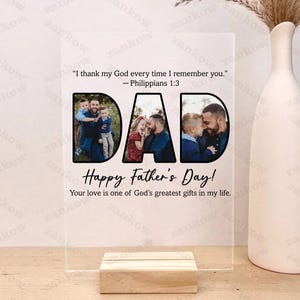 May include: A clear acrylic photo display with the word "DAD" cut out, each letter containing a family photo. The display includes the text "Happy Father's Day!" and a Bible verse. It rests on a wooden base.