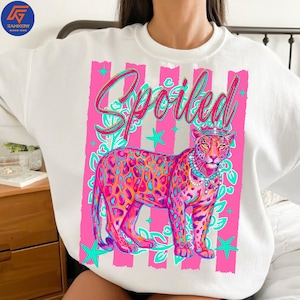 May include: White sweatshirt with a vibrant graphic of a bejeweled leopard wearing a crown, set against a pink striped background. The word "Spoiled" is written in cursive above the leopard.