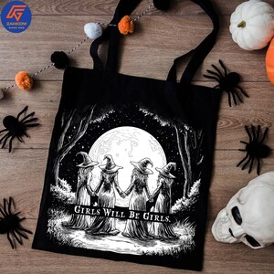 May include: Black tote bag featuring a black and white illustration of four witches holding hands in front of a full moon. The text "Girls Will Be Girls" is below the image. The bag has black handles.
