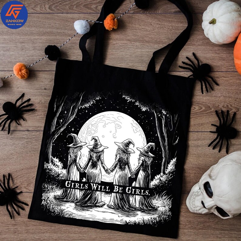 May include: Black tote bag featuring a black and white illustration of four witches holding hands in front of a full moon. The text "Girls Will Be Girls" is below the image. The bag has black handles.