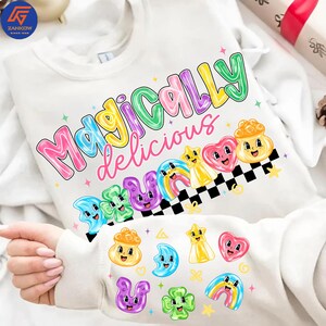 Puede incluir: Sudadera blanca con la frase "Magically Delicious" en letras coloridas. El diseño incluye imágenes de dibujos animados de un arcoíris, estrellas, corazones y otras formas caprichosas. La sudadera tiene cuello redondo y mangas largas.