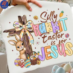May include: White Easter-themed pillow with a cartoon bunny, cross, and the text "Silly Rabbit Easter is for Jesus." The design includes colorful eggs, a basket of carrots, and floral accents. The pillow is held by a person.