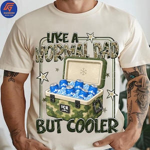 May include: Beige t-shirt with a graphic featuring a camouflage-style text that reads "LIKE A NORMAL DAD BUT COOLER." The graphic includes a green cooler filled with blue cans and ice cubes, and star accents.