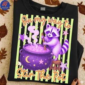May include: A black sweatshirt featuring a graphic of a purple raccoon standing on a small wooden stool, stirring a purple cauldron with a wooden spoon. The design includes light green vertical stripes, pink stars, and the text On my way to Stir The Pot.