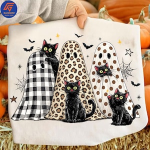 May include: A light-colored t-shirt featuring a graphic of three black cats dressed as ghosts. One ghost has a black and white buffalo check pattern, one has a leopard print, and one has brown polka dots. Small bats, stars, and spiderwebs surround them.