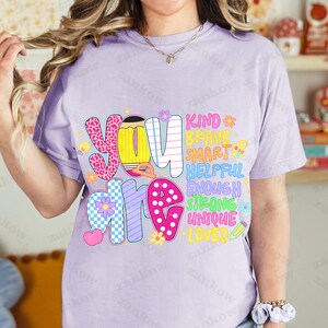 May include: A light purple t-shirt with the words "YOU ARE" in colorful, patterned letters. The words "KIND, BRAVE, SMART, HELPFUL, ENOUGH, STRONG, UNIQUE, LOVED" are printed vertically on the right side of the shirt.
