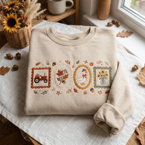 May include: A beige crewneck sweatshirt featuring four embroidered rectangular and oval frames across the chest. The designs include a red tractor with a pumpkin, autumn leaves with an acorn, a goose wearing a hat and scarf, and sunflowers in a vase.