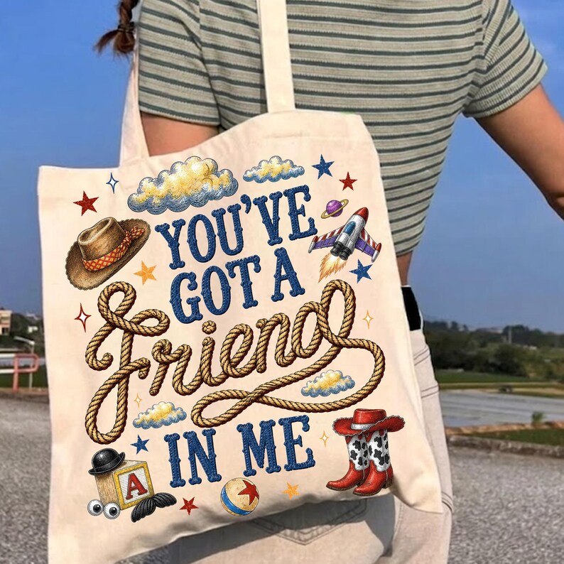 May include: A cream-colored canvas tote bag featuring the blue text "YOU'VE GOT A friend IN ME" written in a mix of block and rope-style fonts. The design includes illustrations of a cowboy hat, a rocket, a block with the letter A, and cowboy boots.