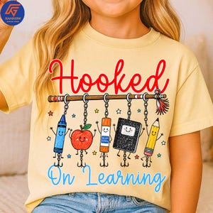 May include: A pale yellow t-shirt featuring a graphic print with the red text Hooked and blue text On Learning. The design shows a fishing rod with hanging school supplies including a blue crayon, red apple, glue stick, notebook, and yellow pencil.