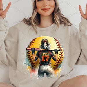 K&ouml;nnte beinhalten: Ein beiger Pullover mit einem Aquarell-Design einer Softballspielerin, die einen Schl&auml;ger h&auml;lt. Der Softball ist gelb mit roten N&auml;hten und die Spielerin tr&auml;gt ein schwarzes Trikot mit dem Namen "Emma" und der Nummer "17" auf dem R&uuml;cken.