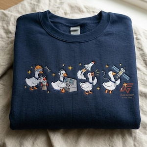 May include: A navy blue crewneck sweatshirt featuring an embroidered design of four white geese in space-themed roles. The geese are depicted wearing a hard hat, using a computer, launching a rocket, and operating a satellite. The text ZANKIW is visible.