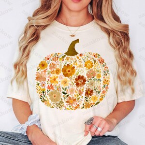 May include: Cream-colored t-shirt featuring a floral pumpkin design. The pumpkin is composed of various flowers in shades of orange, yellow, and brown, with a green stem. A fall-themed graphic tee for autumn.