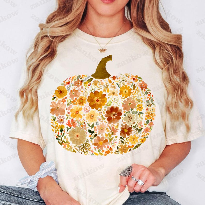 May include: Cream-colored t-shirt featuring a floral pumpkin design. The pumpkin is composed of various flowers in shades of orange, yellow, and brown, with a green stem. A fall-themed graphic tee for autumn.