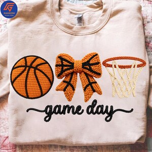 May include: Cream-colored sweatshirt with a basketball, bow, and hoop design. The basketball and bow are orange with black accents. The text "game day" is in black script. A fun, sporty design.