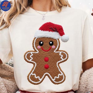 Peut inclure: T-shirt crème avec un motif de bonhomme en pain d'épices. Le bonhomme en pain d'épices a un corps marron, des bordures en glaçage blanc et un bonnet de Père Noël rouge. Le motif comprend des yeux noirs, un sourire rose et des boutons rouges. Article de fête.