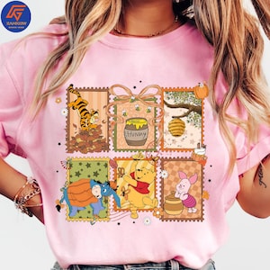 May include: Pale pink t-shirt with a colourful graphic of Winnie the Pooh characters in postage stamp designs. The design includes Tigger, Eeyore, Winnie the Pooh, Piglet, a honey pot, and a beehive. The word "Hunny" is also visible.