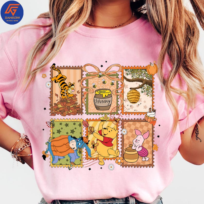 May include: Pale pink t-shirt with a colourful graphic of Winnie the Pooh characters in postage stamp designs. The design includes Tigger, Eeyore, Winnie the Pooh, Piglet, a honey pot, and a beehive. The word "Hunny" is also visible.