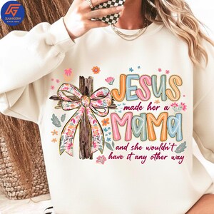 May include: A cream-coloured sweatshirt with a colourful graphic. The design features a wooden cross with a floral bow, the word "JESUS" in orange and blue, and the word "MAMA" in pink and blue, with the phrase "and she wouldn't have it any other way."