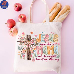 May include: A cream tote bag with the text "JESUS made her a MAMA and she wouldn't have it any other way." The bag features a cross with a floral bow. Apples and bread are also in the image.