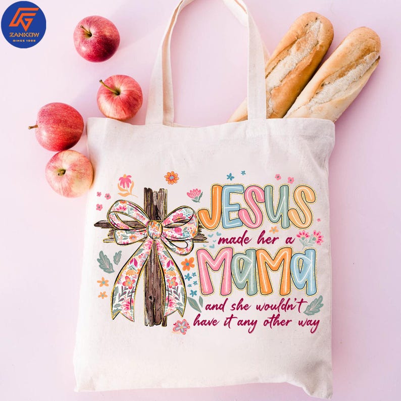 May include: A cream tote bag with the text "JESUS made her a MAMA and she wouldn't have it any other way." The bag features a cross with a floral bow. Apples and bread are also in the image.