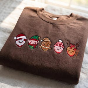 May include: A brown crewneck sweatshirt featuring five embroidered holiday-themed anatomical heart characters. The designs include a Santa heart, an elf heart, a gingerbread heart, a heart in a winter hat, and a reindeer heart with antlers.