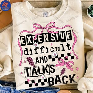 Puede incluir: Una sudadera de color crema con un estampado gráfico en negro y rosa que dice "Caro, difícil y habla mal". El gráfico incluye lazos rosas, estrellas y un patrón a cuadros.