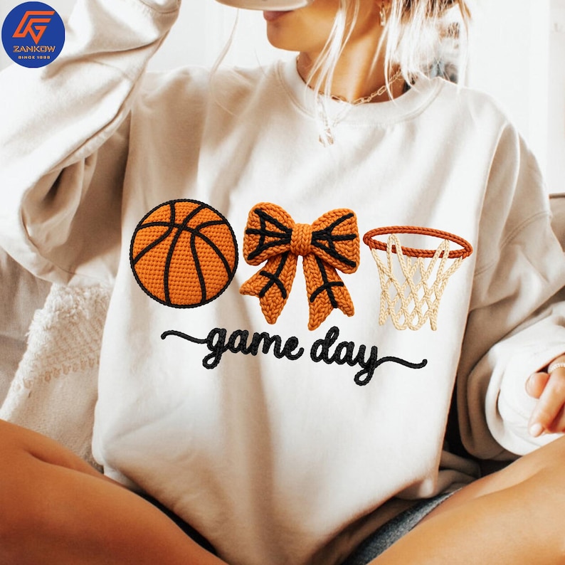 May include: Cream-colored sweatshirt with a basketball, bow, and hoop design. The basketball and bow are orange with black accents. The text "game day" is written in a cursive font below the graphics. The sweatshirt has a crew neck.