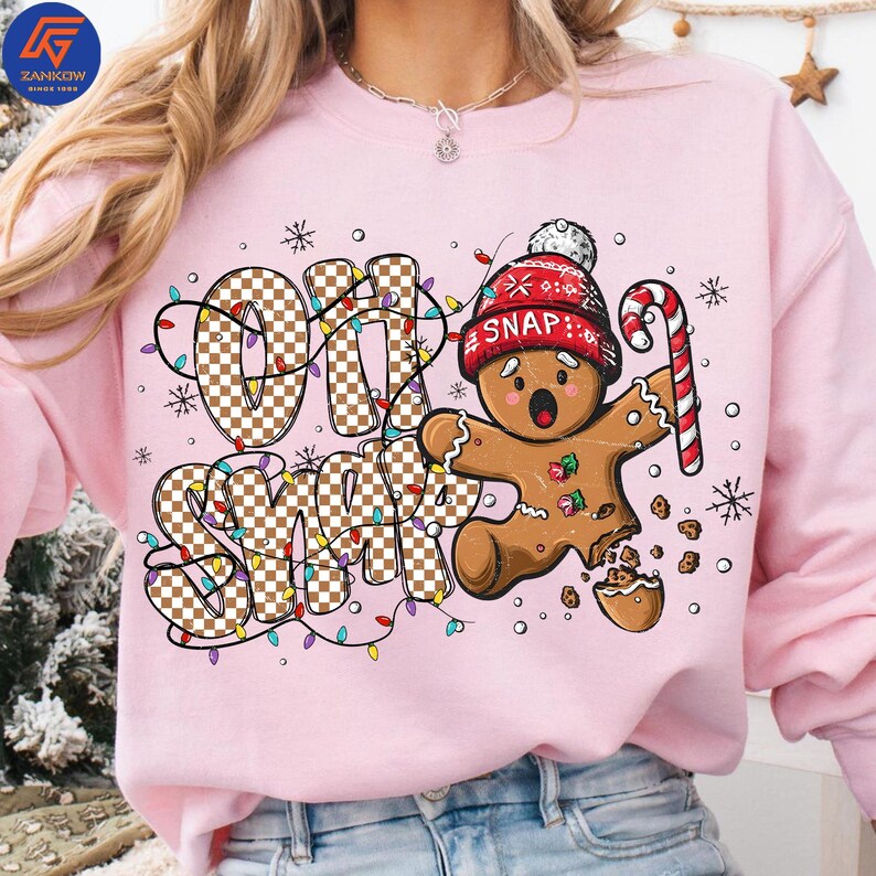 May include: Pink sweatshirt with the text "Oh Snap" in a checkered pattern, decorated with Christmas lights and snowflakes. A gingerbread man wearing a red hat and holding a candy cane is featured in the design.
