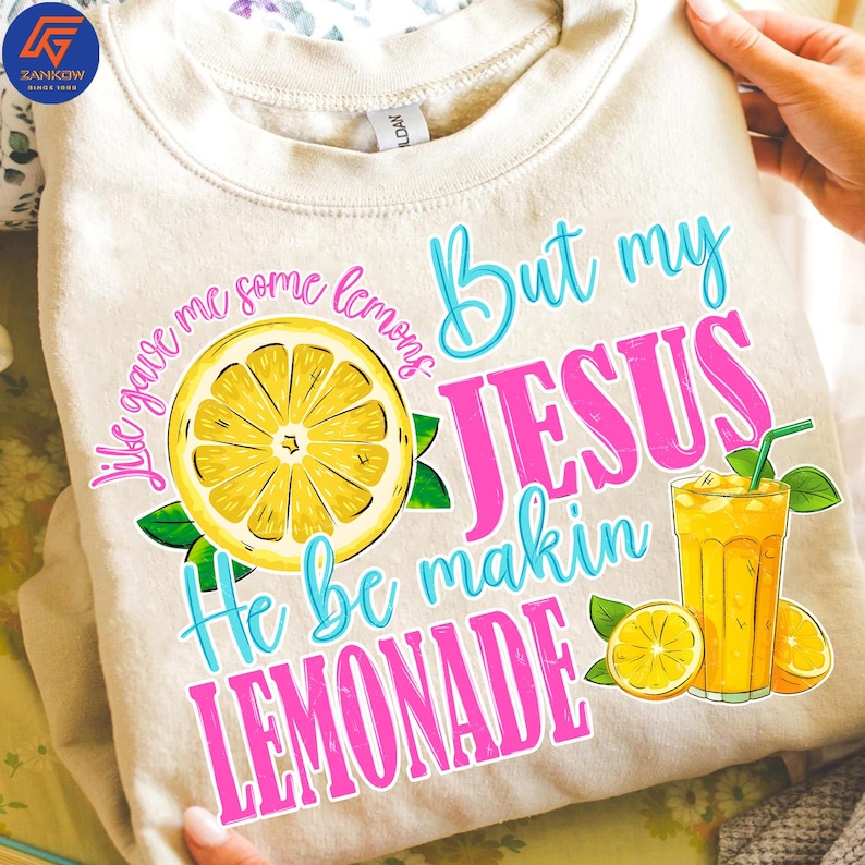 May include: Beige sweatshirt with a colorful graphic. The design features a large yellow lemon slice, a glass of lemonade, and the text "Life gave me some lemons, But my JESUS be makin LEMONADE" in various colors.