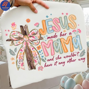 May include: White t-shirt with the text "JESUS made her a MAMA and she wouldn't have it any other way." The shirt features a wooden cross with a floral bow and colourful floral accents.